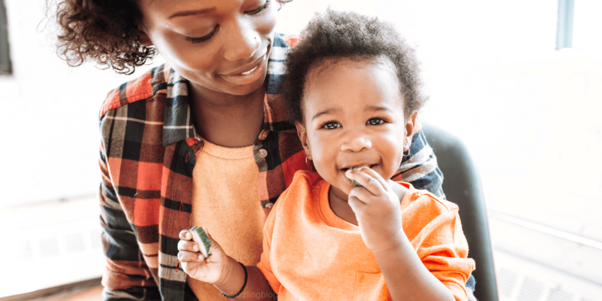 Black mother feeding her baby cucumbers. what to buy your toddler blog post banner