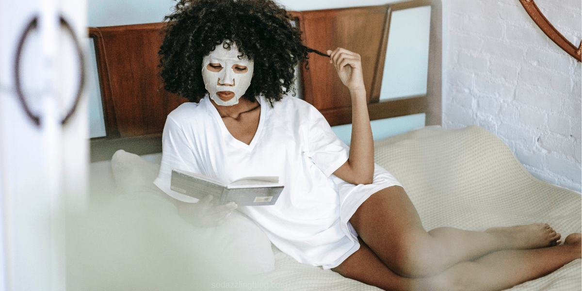 Black woman with a facial sheet mask relaxing on a bed while reading a book. She is enjoying some time to herself as she practices self-care a stay at home mom