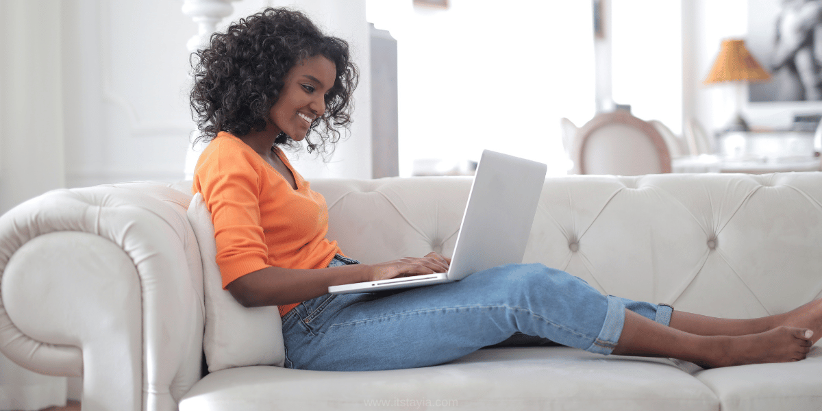 black woman wearing blue jeans and orange shirt sitting on a couch with a laptop researching tips for growth on YouTube